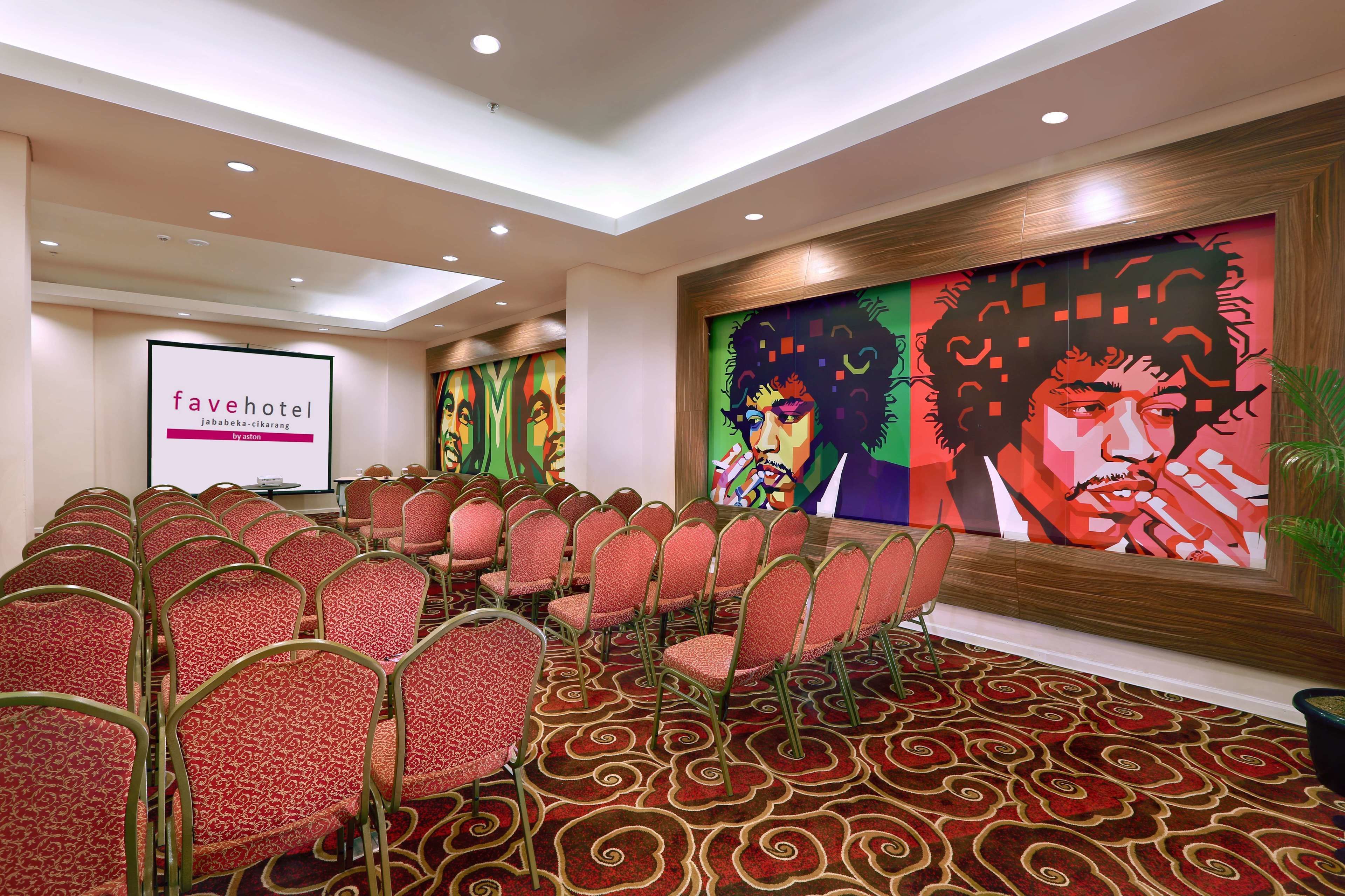 A conference room with rows of red chairs, a projector screen displaying 'favehotel', and a large abstract painting on the wall.