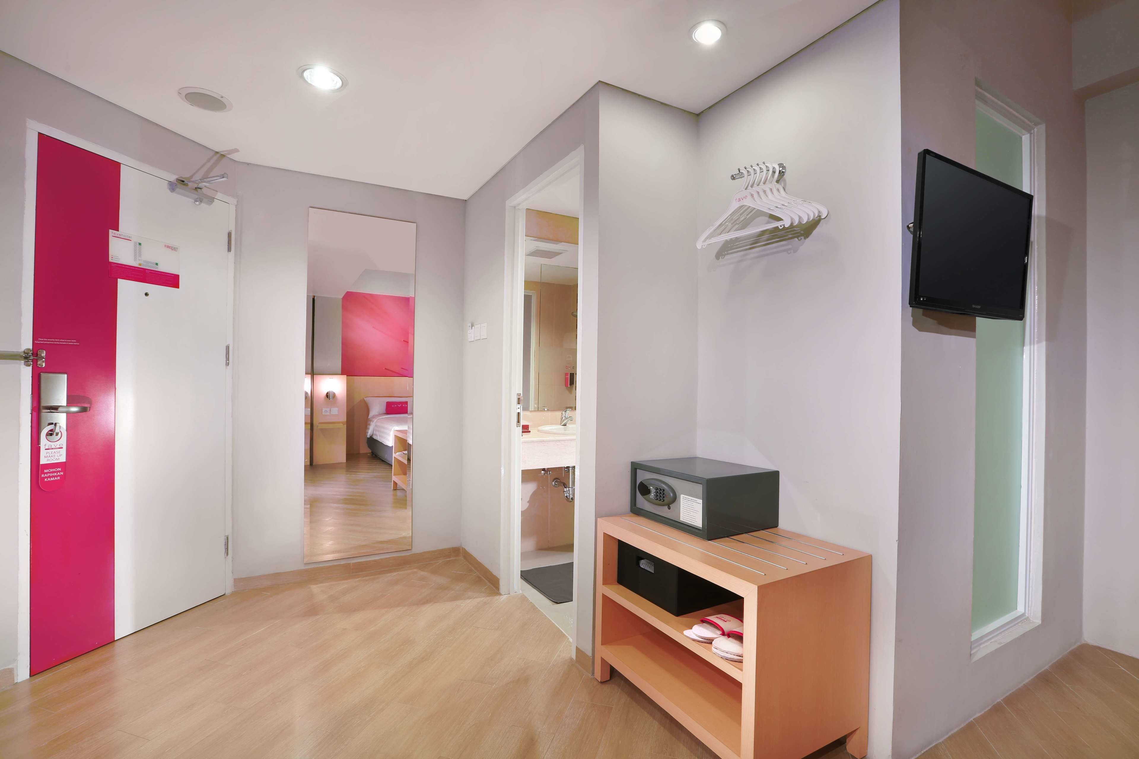 Un couloir d’hôtel avec un plancher en bois, une porte rose, une porte blanche, un miroir, un téléviseur fixé au mur et une petite armoire en bois sur laquelle est posé un four à micro-ondes.