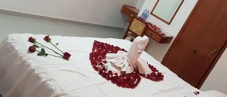 Ein Schlafzimmer mit einem Bett, das mit weißen Laken und Rosenblüten bedeckt ist. Auf dem Bett liegen eine Fernbedienung und ein Smartphone. Ein Nachttisch aus Holz mit einem Spiegel und ein kleiner Tisch mit einer Wasserflasche stehen bereit.