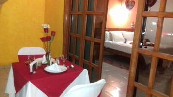 Un comedor con una mesa preparada para dos, un florero con flores y vista a una cama decorada con forma de corazón.