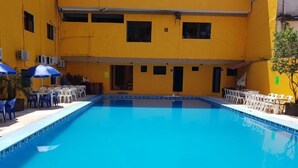A swimming pool with blue tiles, surrounded by white chairs and umbrellas, and a yellow building with multiple windows and doors. - Hotel Arcos del Parque (Acayucan)