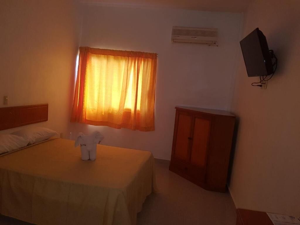 Habitación de hotel con cama, una mesita con un osito de peluche, un armario de madera y un televisor fijado a la pared.