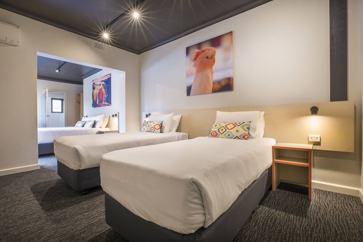 A hotel room with two beds, a nightstand, a wall-mounted lamp, and a painting of a bird.