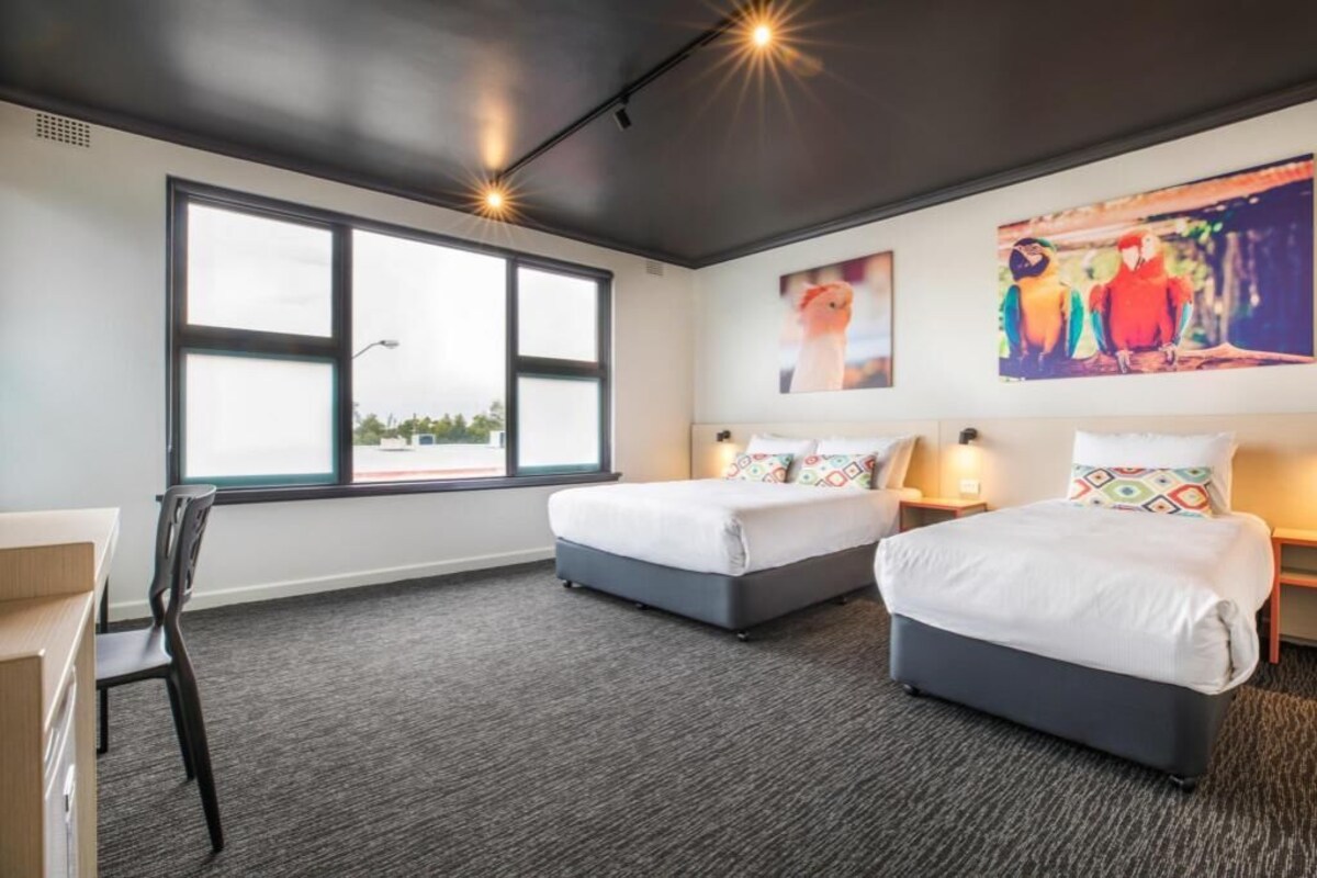 Une chambre d’hôtel avec deux lits, un bureau, une chaise, une grande fenêtre et des œuvres d’art accrochées au mur.