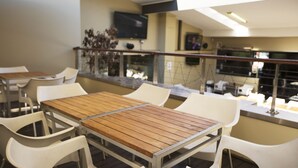 Restaurante com mesas de madeira e cadeiras brancas, uma TV na parede e um corrimão com vista para a rua.