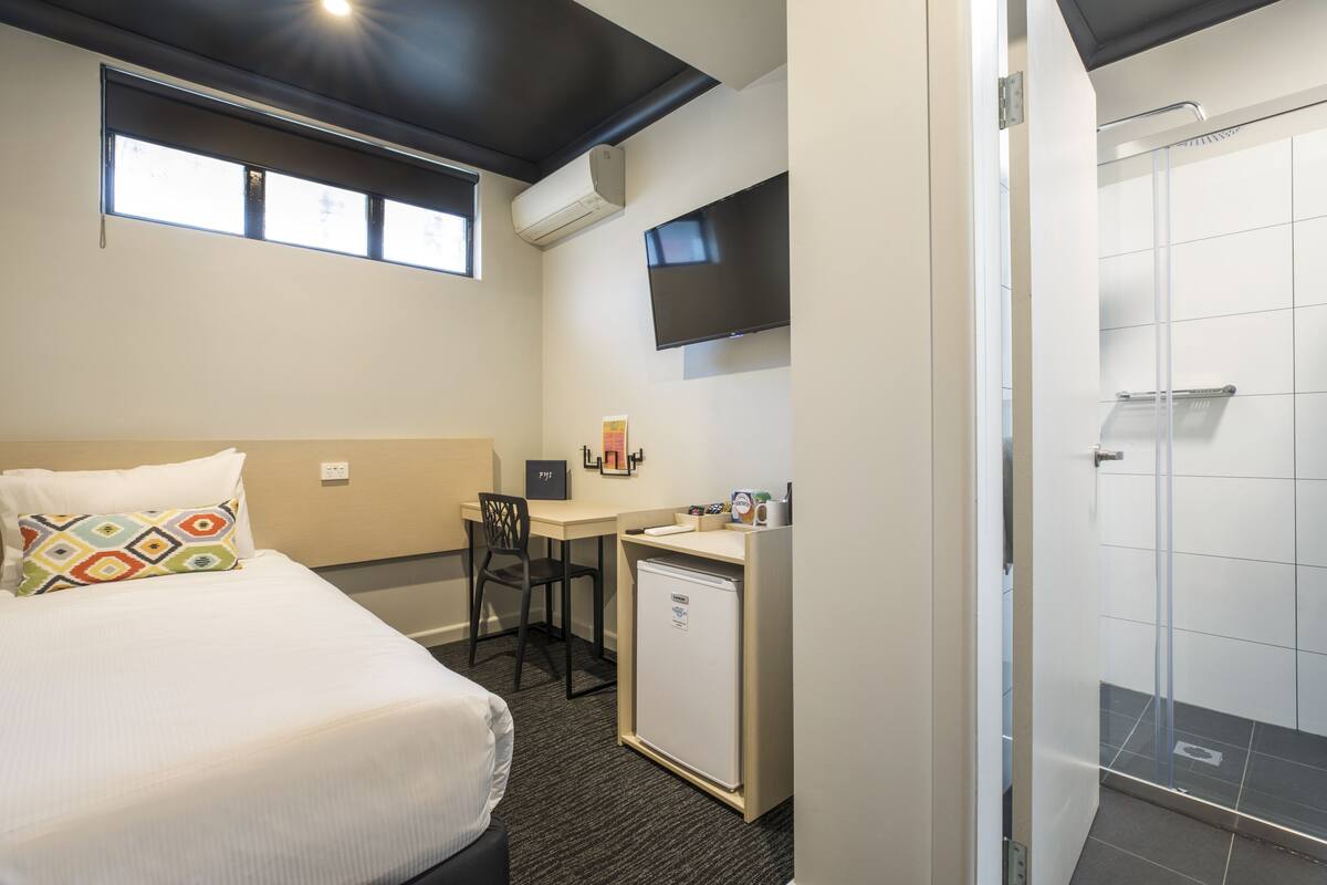 A hotel room with a bed, desk, chair, small refrigerator, TV, and a shower area.