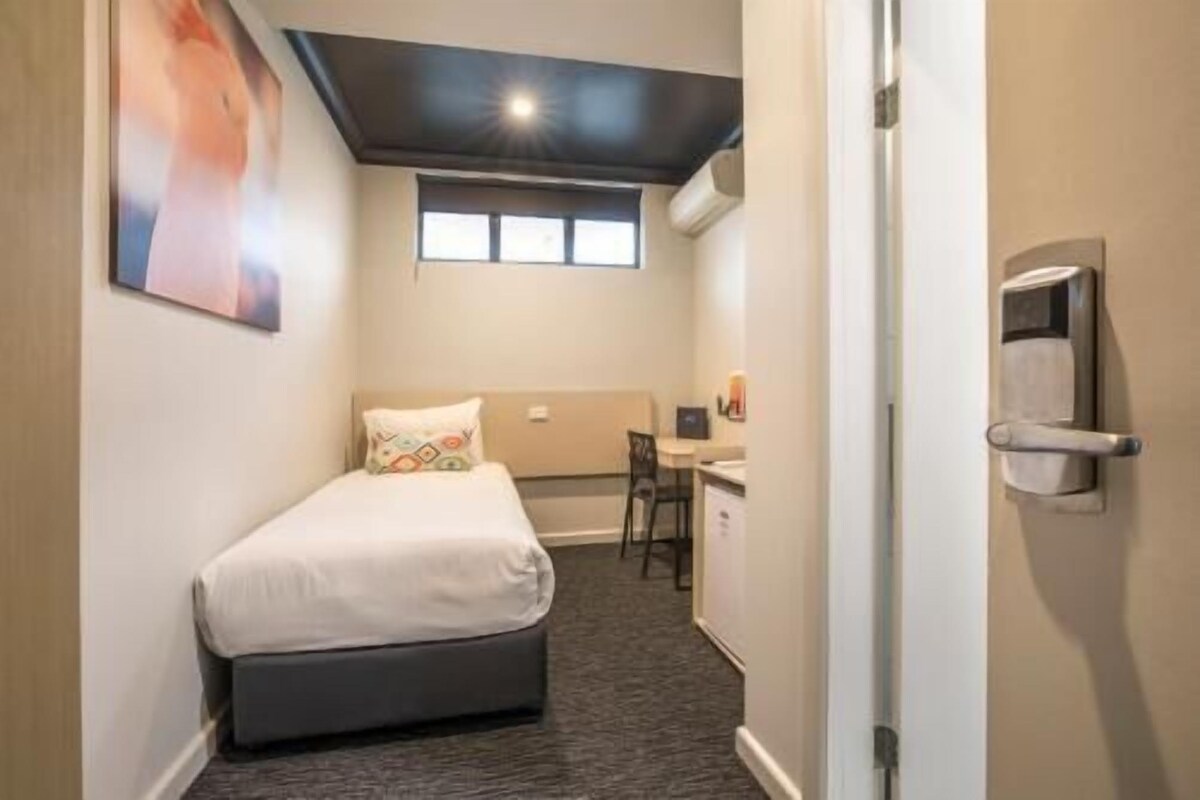 Une chambre d’hôtel de taille réduite, comprenant un lit simple, un petit bureau, une chaise et une fenêtre.