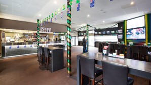 Uma área de bar com diversas opções de assento, um balcão com cardápio e uma televisão exibindo um evento esportivo.