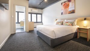 Quarto de hotel moderno com cama grande, mesa de cabeceira, ar condicionado fixado na parede e janela que proporciona luz natural.