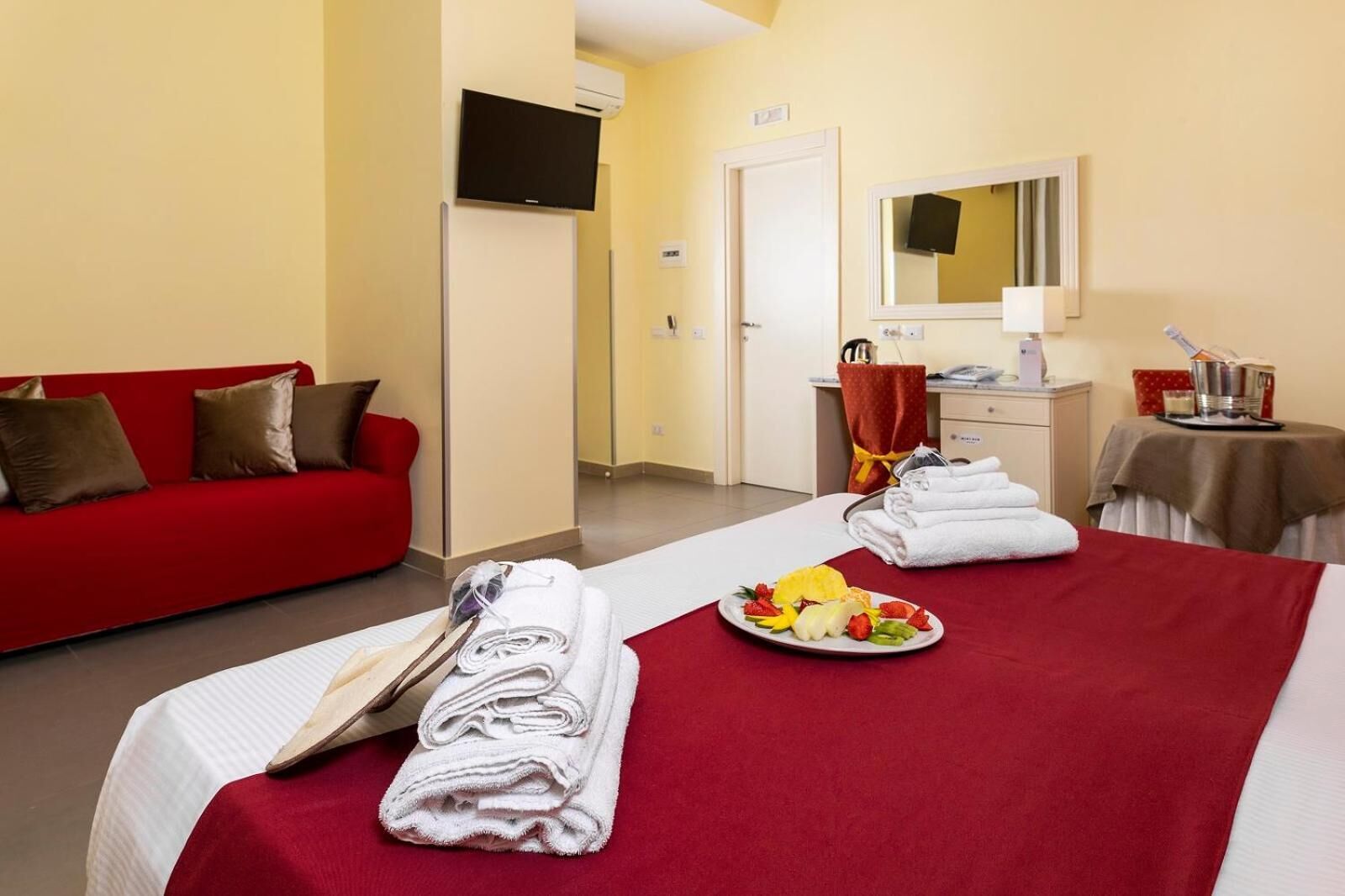 Ett hotellrum med en röd soffa, en säng med ett rött överkast, en tallrik med frukt och handdukar.