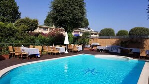 En swimmingpool med solstolar och bord, omgiven av ett trädäck och träd.