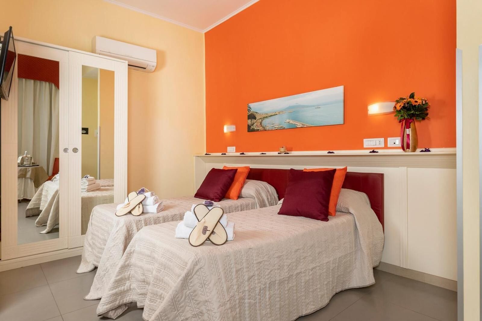 Camera d'albergo con due letti, un grande specchio, un condizionatore d'aria, un quadro raffigurante una scena costiera e un vaso con fiori sul comodino.