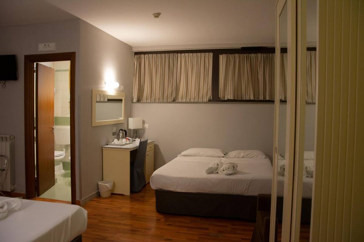 A hotel room with a wooden floor, a bed with white linens, a bedside table with a lamp, a small desk with a chair, and a bathroom visible through an open door.