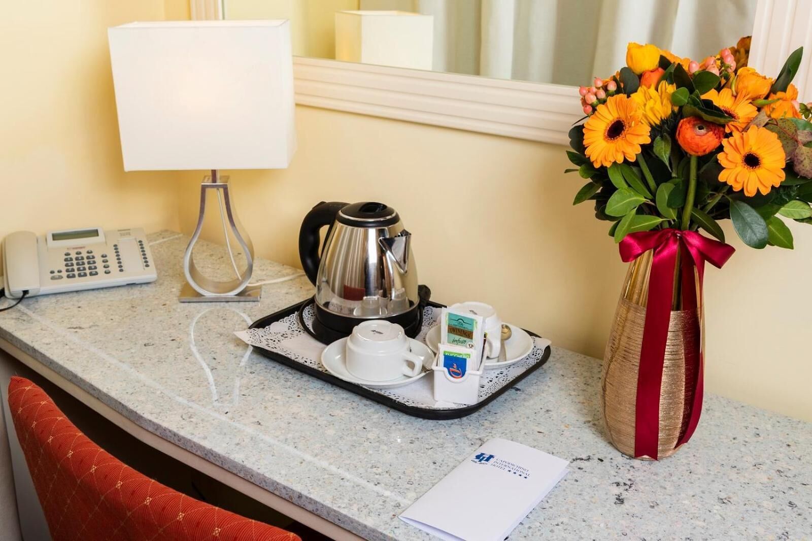 Ein Hotelzimmer-Tresen mit einer Kaffeemaschine, einem Telefon und einem Tablett mit Tassen und einer Taschentuchbox. Eine Vase mit Blumen steht rechts.