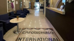 En hotellobby med en receptionsdisk, en sittgrupp och ett dekorativt mosaikgolv med texten ”CAPODICHINO INTERNATIONAL”.