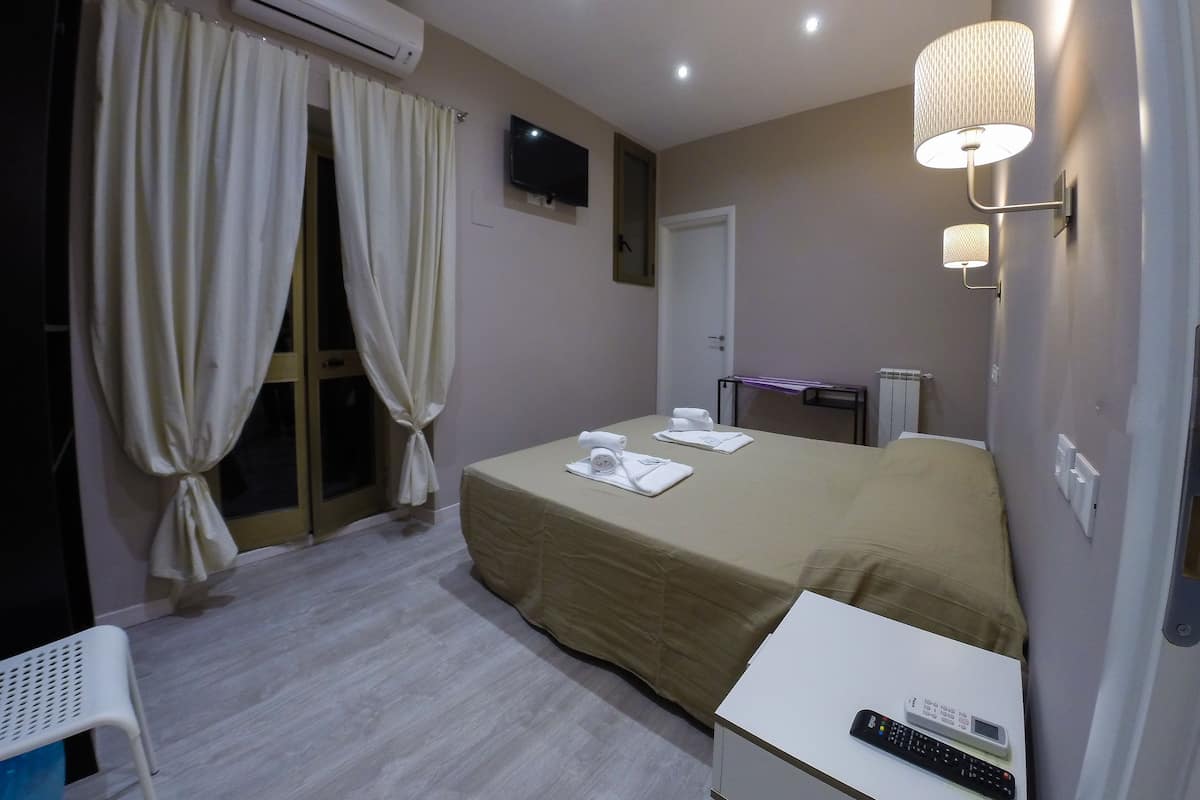 Una camera d'albergo con un letto, una scrivania, una sedia, un condizionatore d'aria, una televisione e una lampada.