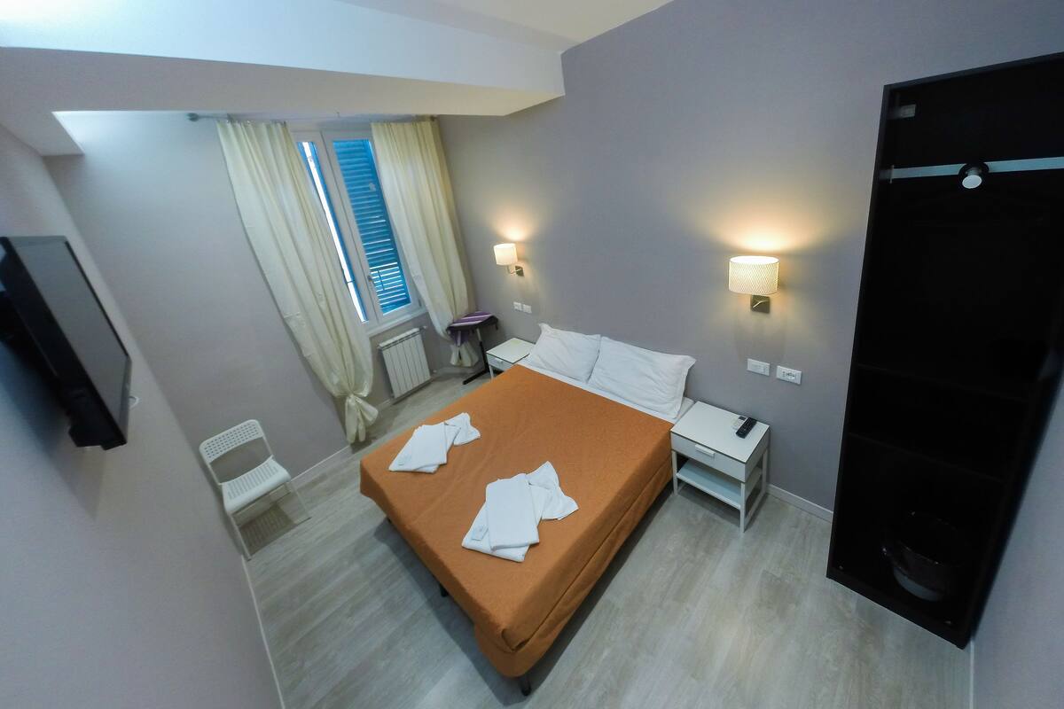 Una camera d'albergo con un letto, comodini, una scrivania, una sedia, una finestra con tende e un lampadario a parete.
