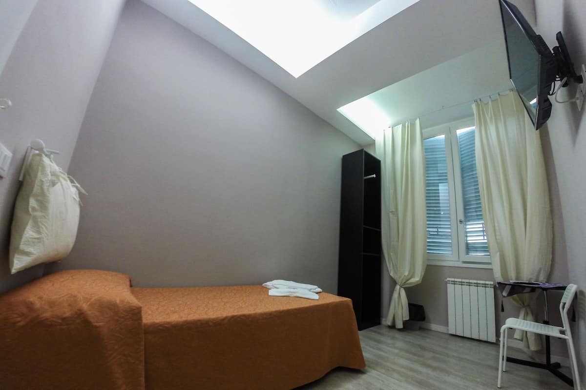 Una piccola camera con un letto singolo, soffitto spiovente, termosifone, finestra con tende e TV a parete.