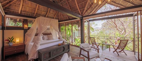 A room with a wooden ceiling, a four-poster bed, a nightstand, a bedside table with a lamp, and a view of trees through large windows.