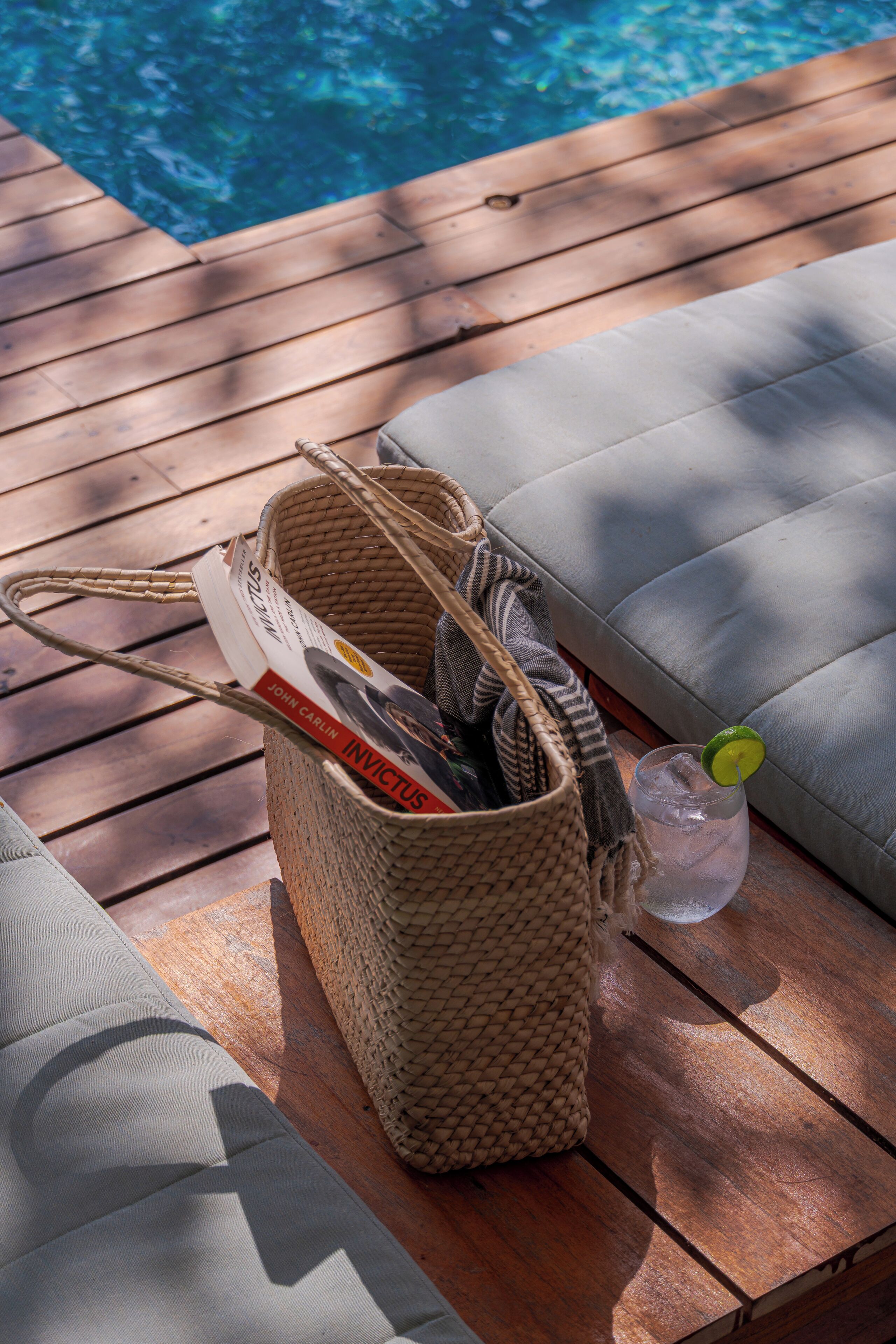 Una terraza de madera con una cesta tejida que contiene un libro y una bebida adornada con una rodaja de lima.