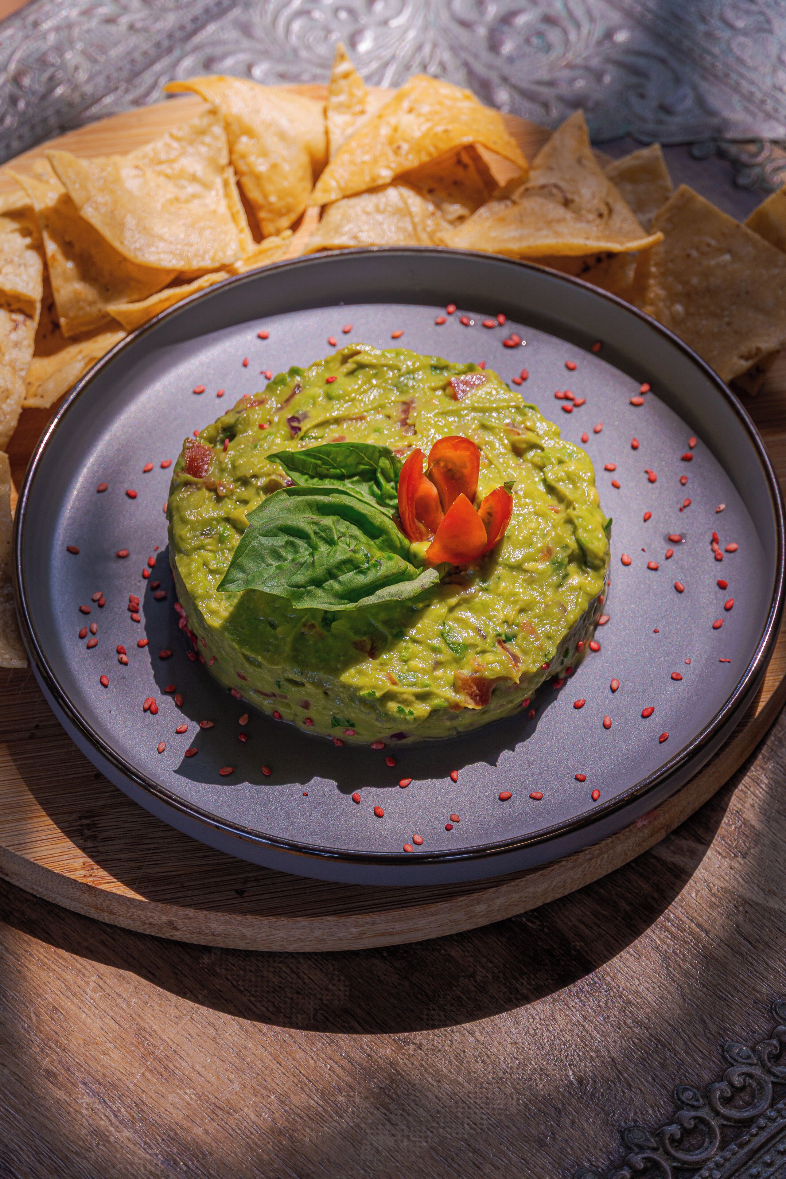 A plate of guacamole with a decorative leaf and tomato on a dark plate, surrounded by tortilla chips.