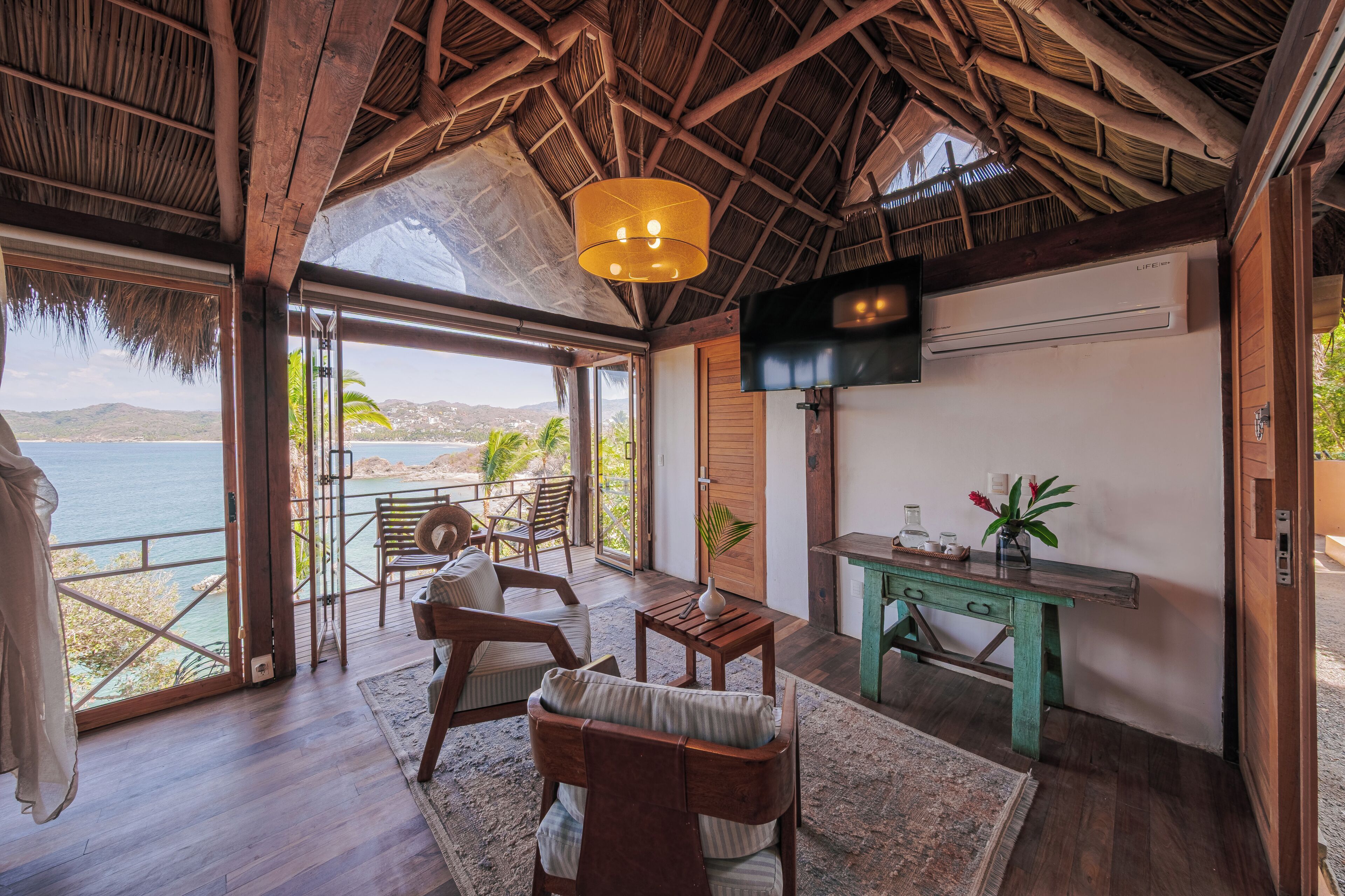 A room with a wooden ceiling, a TV mounted on the wall, a wooden desk, a chair, and a view of the sea.