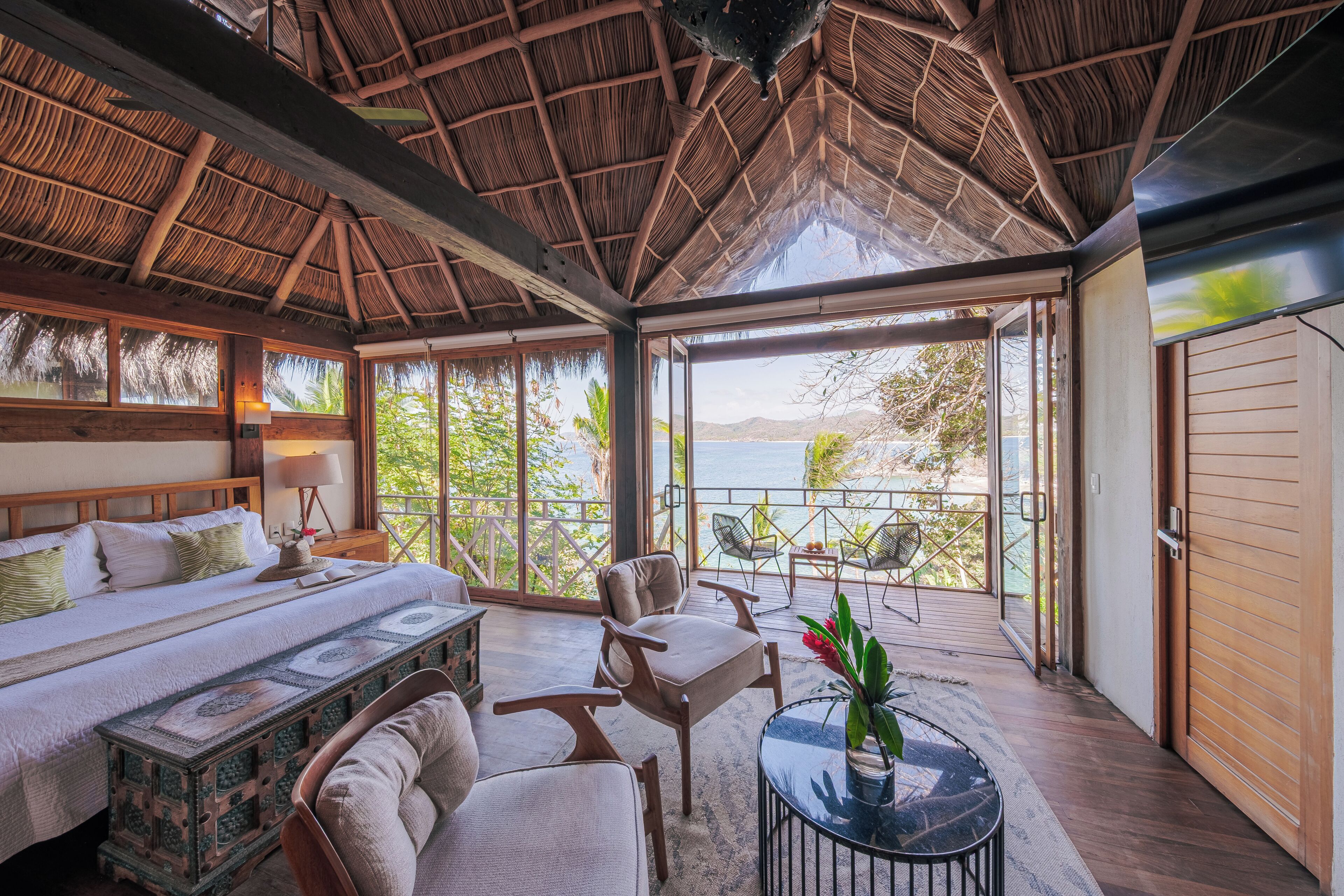 Una habitación con cama, zona de estar con mesa y sillas, un ventanal con vistas al mar y un techo de paja.