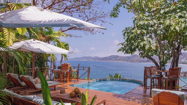 Zona de piscina con sillas reclinables, sombrillas y vistas al mar.
