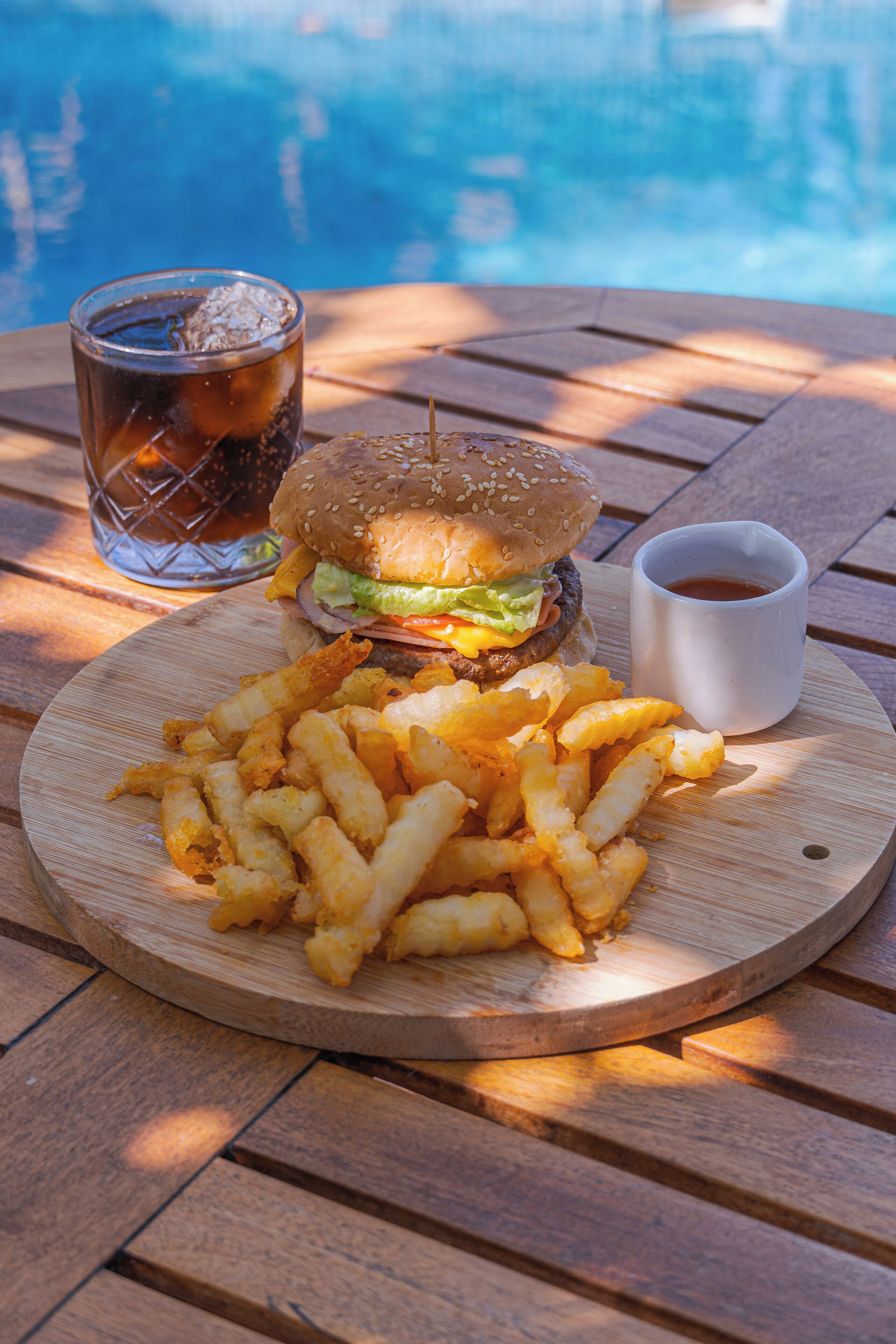 Un repas composé d’un hamburger, de frites et d’une boisson, servi sur une table en bois au bord d’une piscine.