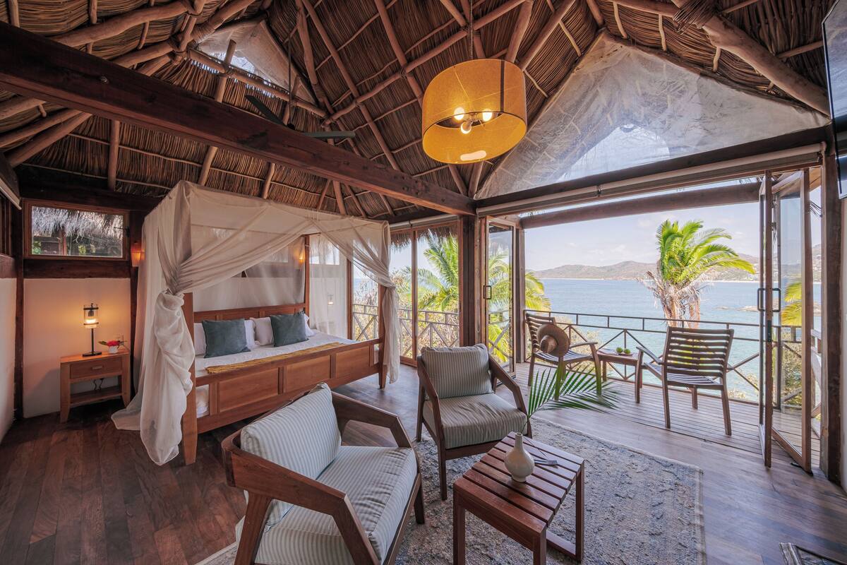 Una habitación con cama, un banco de madera, una silla, una mesa y un ventanal con vistas al mar.