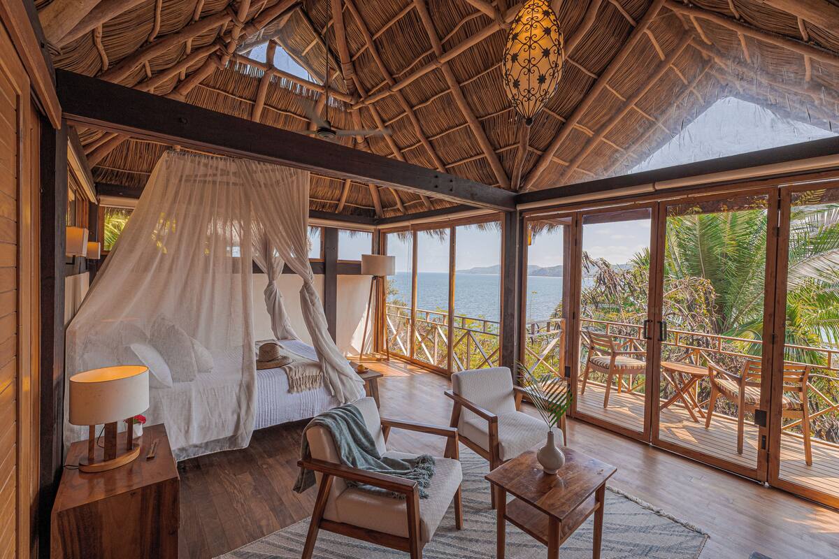 Una habitación con cama, una mesa, sillas, una lámpara y vista al océano.
