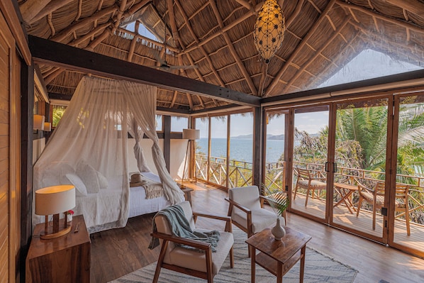 A room with a bed, a table, chairs, a lamp, and a view of the ocean.