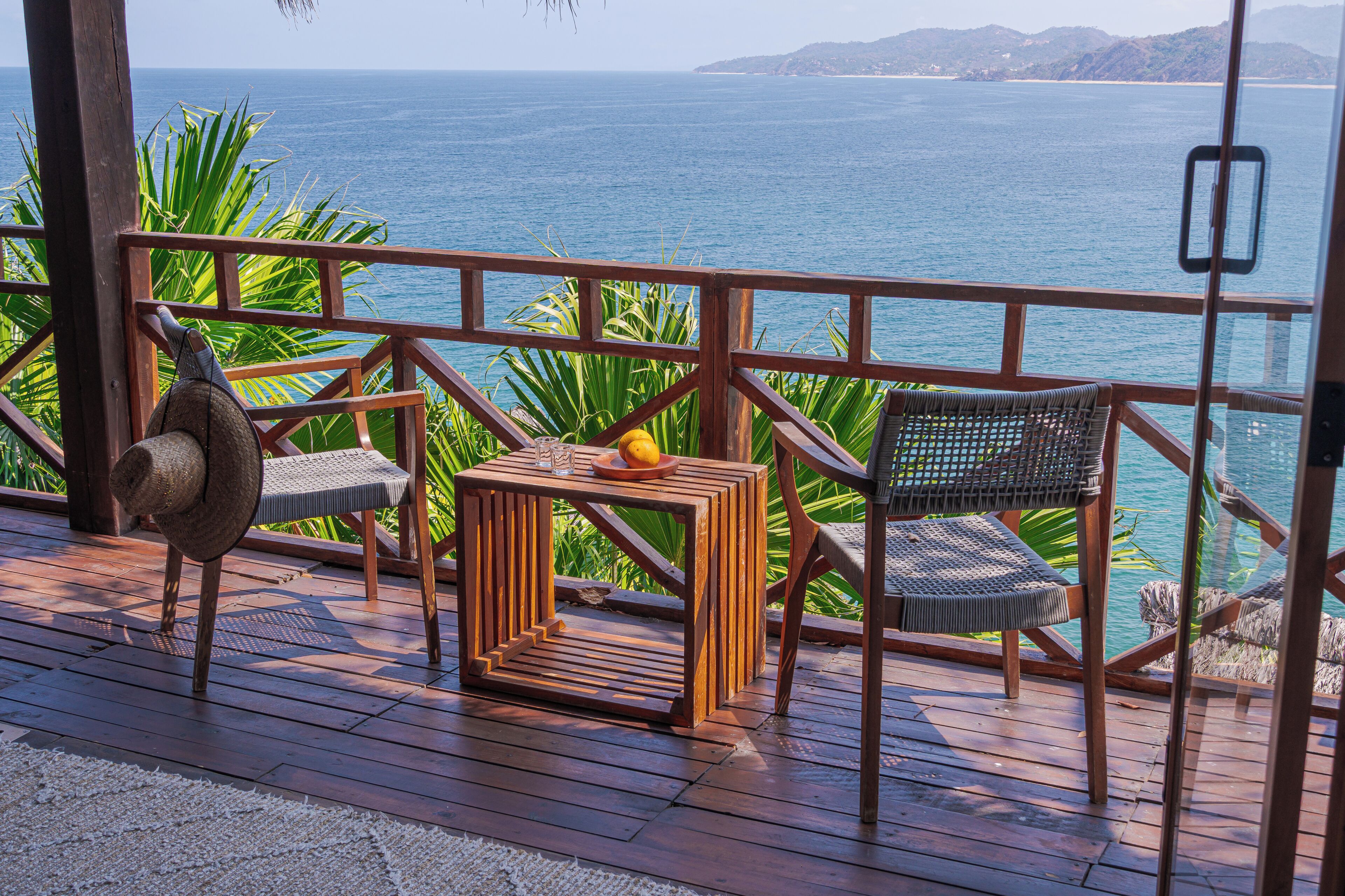 A wooden deck with a view of the ocean, a woven chair, a small table with a fruit, and a woven hat.