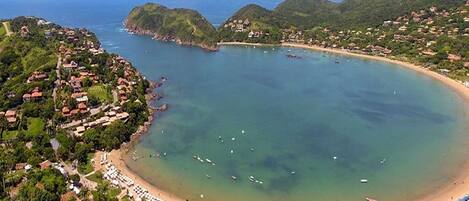 Vista aérea de un pueblo costero con playa, aguas cristalinas y colinas circundantes.