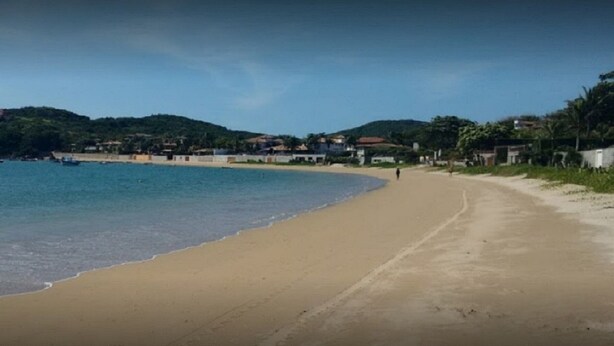 Una playa con orilla arenosa, agua tranquila y un fondo montañoso con edificios.