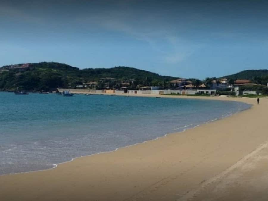 A beach with a sandy shore, calm water, and a hilly background with buildings.