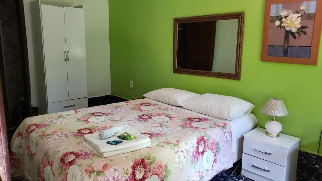 A bedroom with a floral bedspread, a white bedside table with a lamp, a mirror on the wall, and a wardrobe.