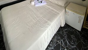 Habitación de hotel con cama, mesita de noche, espejo, refrigerador pequeño y un cuadro en la pared.