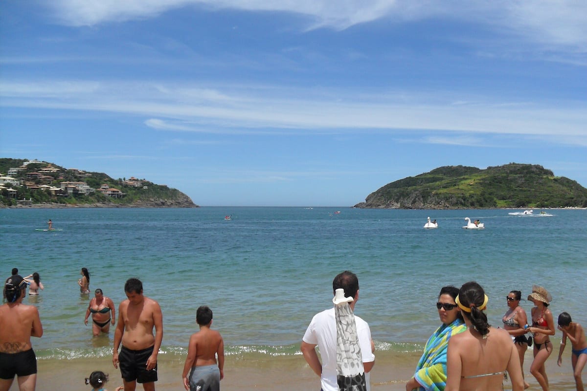 A beach scene with people swimming and standing in the water, boats in the distance, and hills in the background.