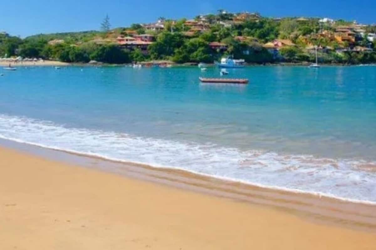 A beach with clear blue water, a boat, and houses on a hill in the background.
