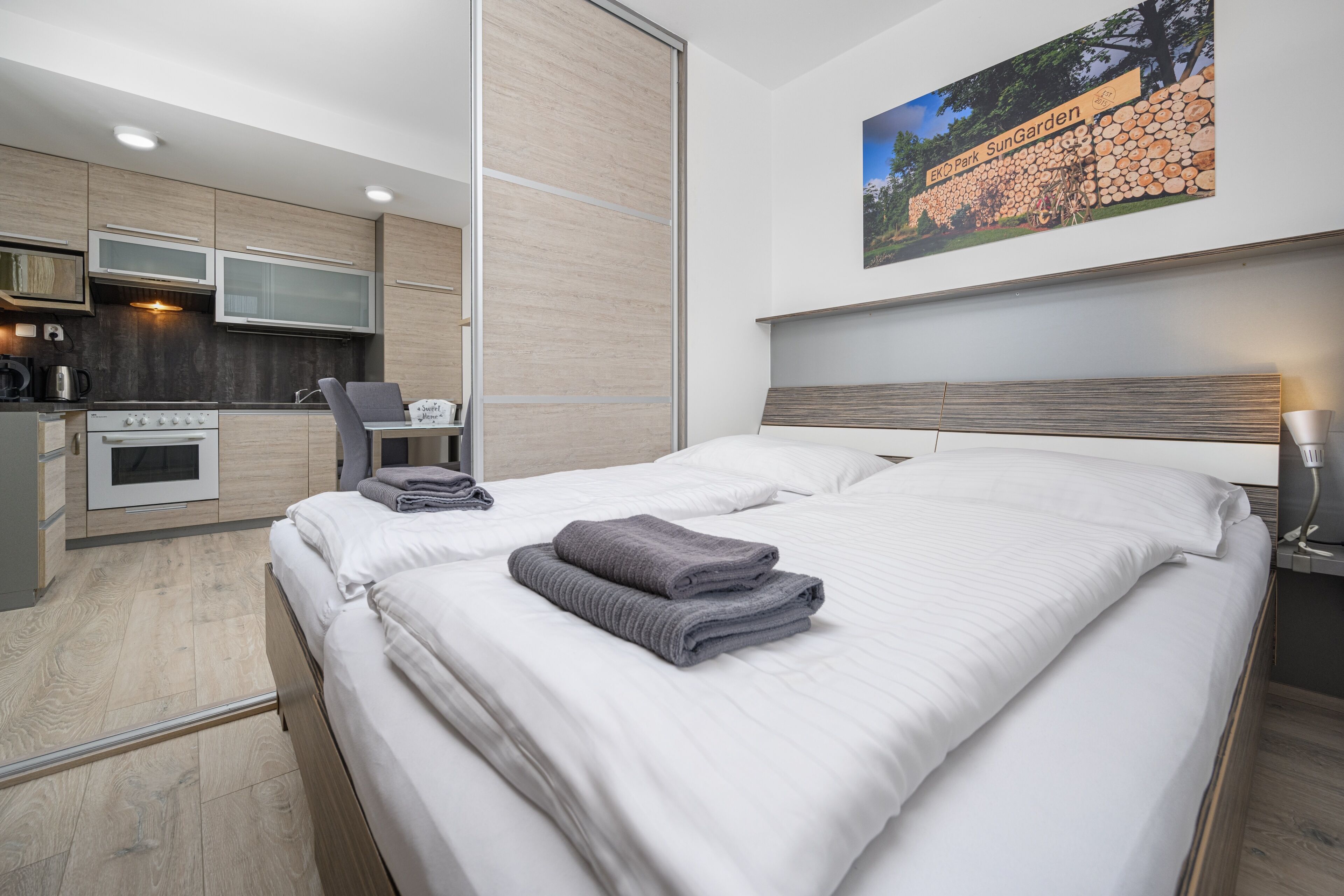 Une chambre d’hôtel moderne équipée d’un lit, d’un bureau, d’une chaise, d’une kitchenette et ornée d’un tableau représentant un parc.