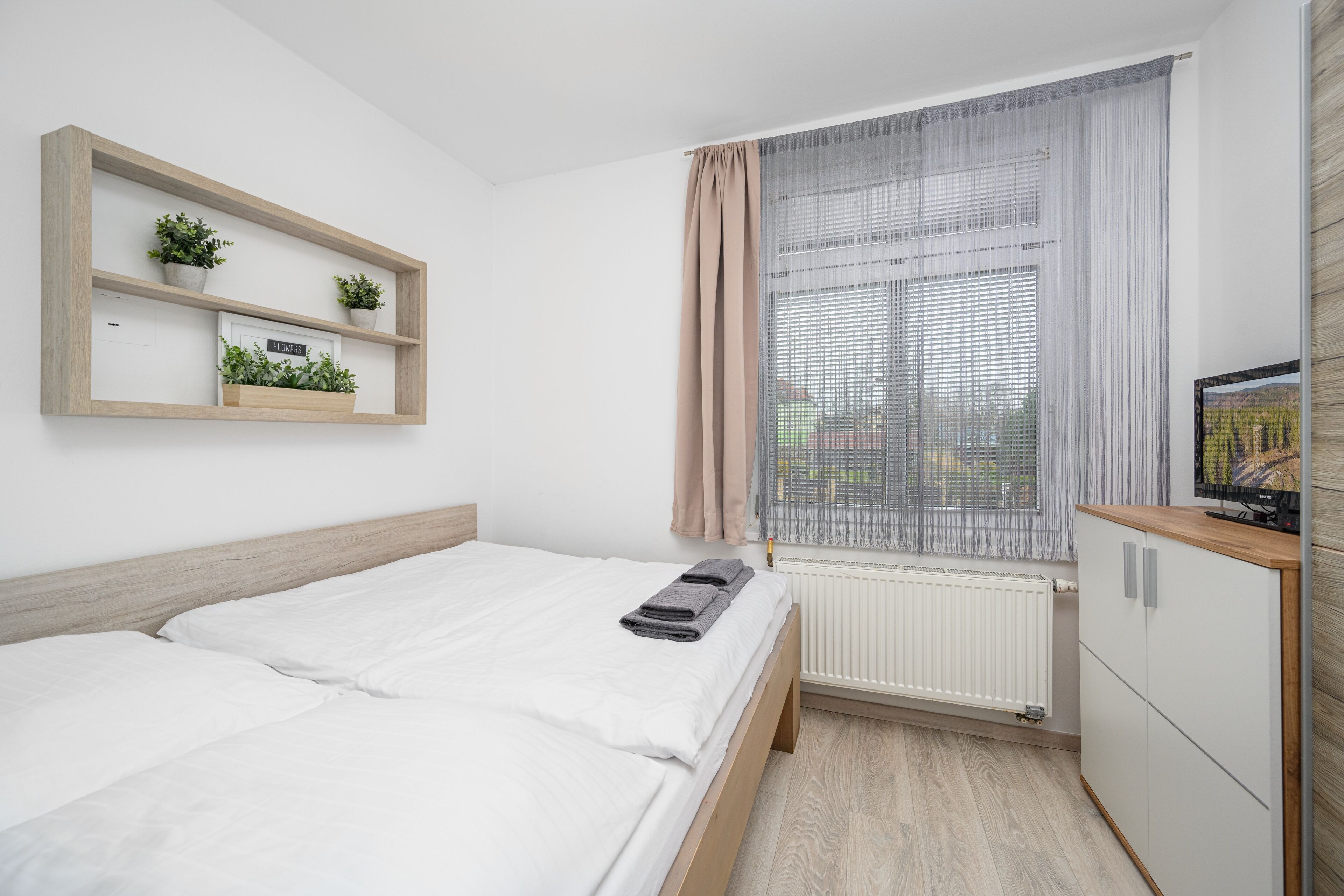 Un dormitorio con una cama, un radiador, una ventana con cortinas, una estantería con plantas y un televisor sobre un mueble.