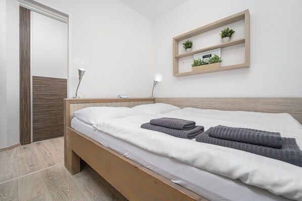 A modern bedroom with a wooden bed, white bedding, a bedside table with a lamp, and a wall-mounted shelf with plants.
