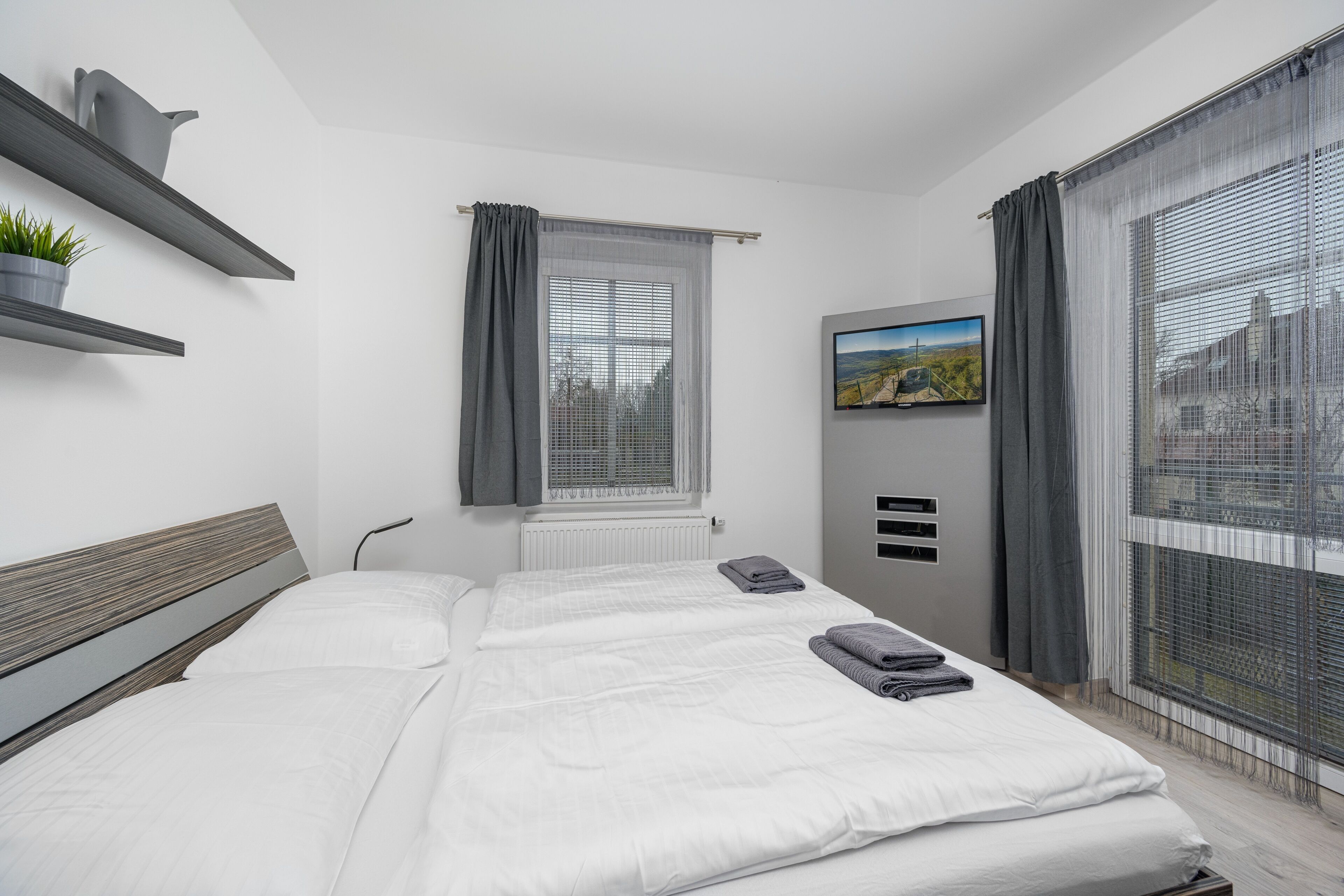 A modern bedroom with a bed, a TV, a window with curtains, and a plant on a shelf.