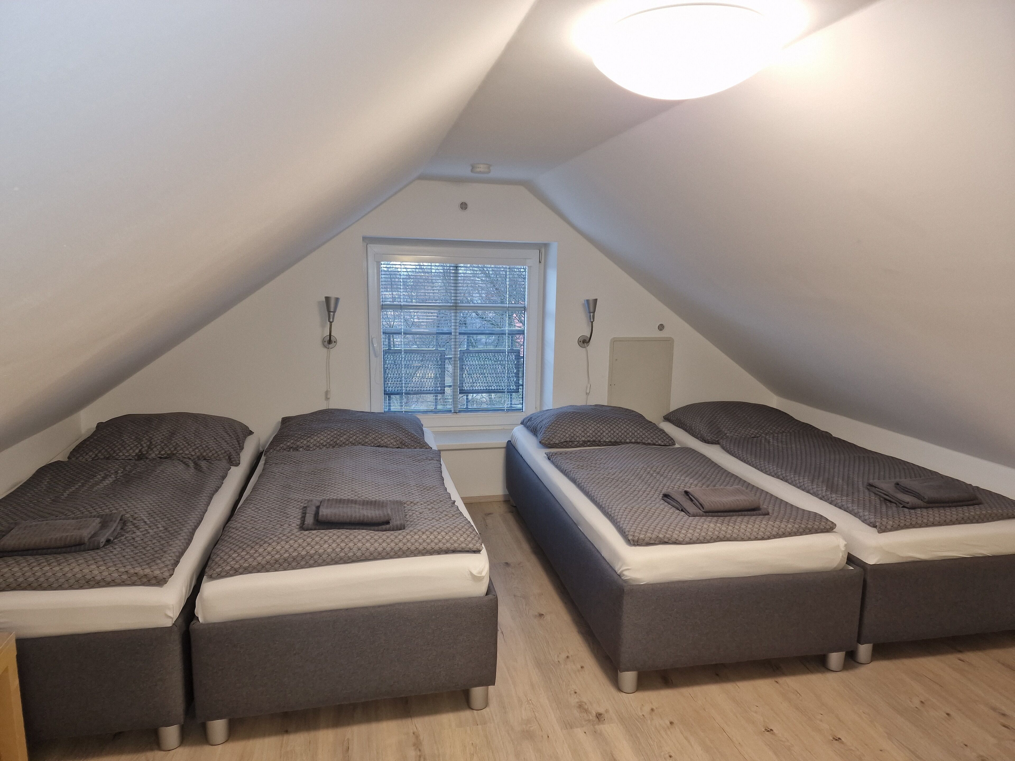 A dormitory room with four single beds arranged in a U-shape, a window with blinds, and a ceiling light.