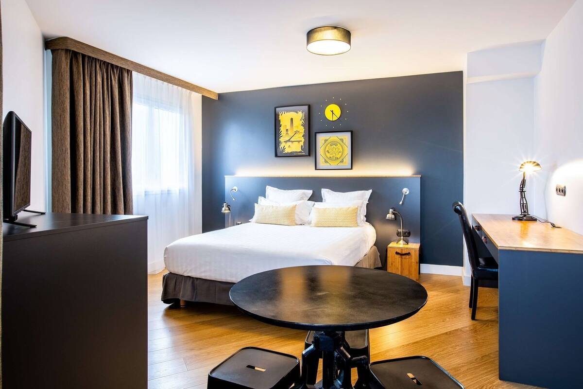 A modern hotel room with a bed, bedside tables, a desk, and a round table.