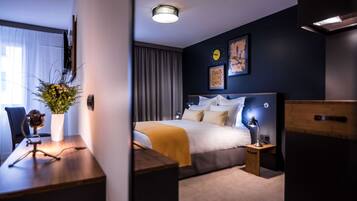 Una habitación de hotel moderna con una cama bien hecha, un escritorio con una computadora portátil, una pequeña mesa con un florero y un ventanal con cortinas.