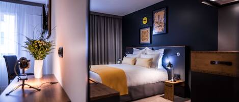 A modern hotel room with a neatly made bed, a desk with a laptop, a small table with a vase of flowers, and a large window with curtains.