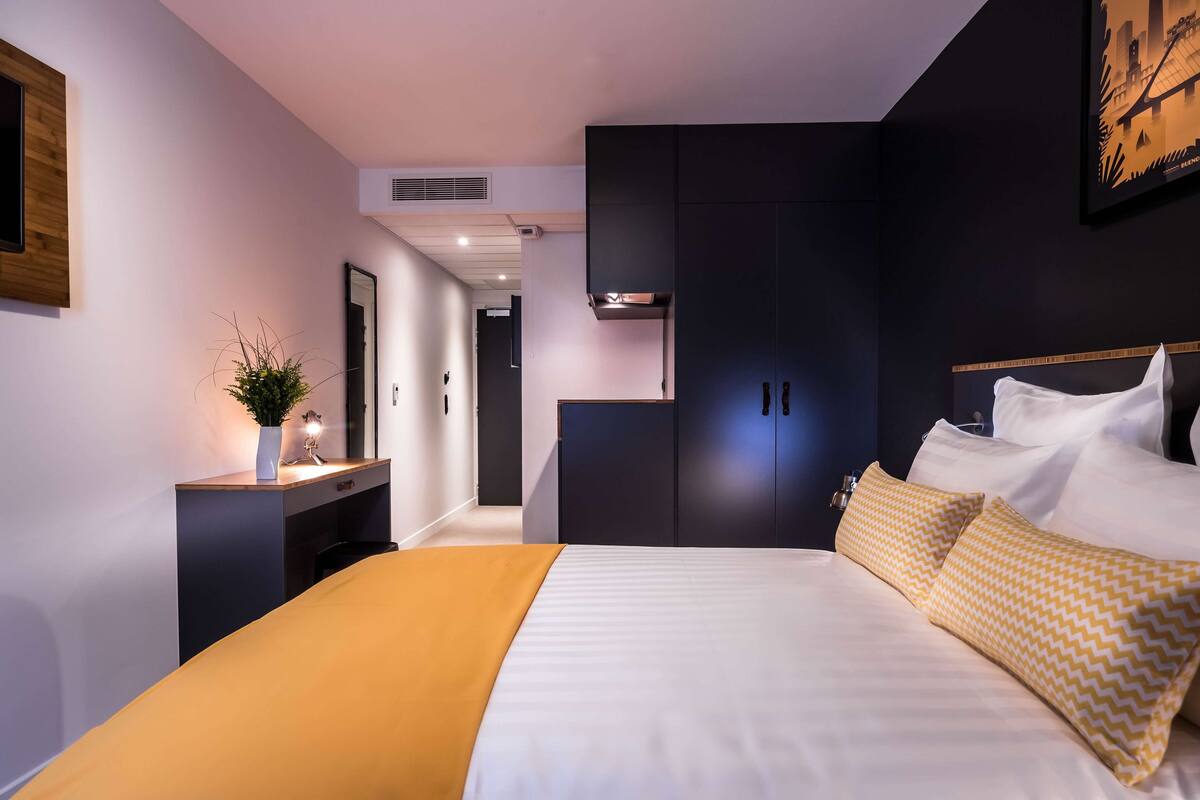 A modern hotel room with a large bed, a bedside table with a lamp, a dark wardrobe, and a framed picture on the wall.