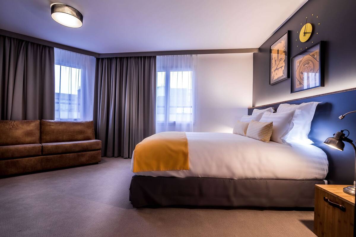 A modern hotel room with a large bed, a sofa, a bedside table, and a wall clock.